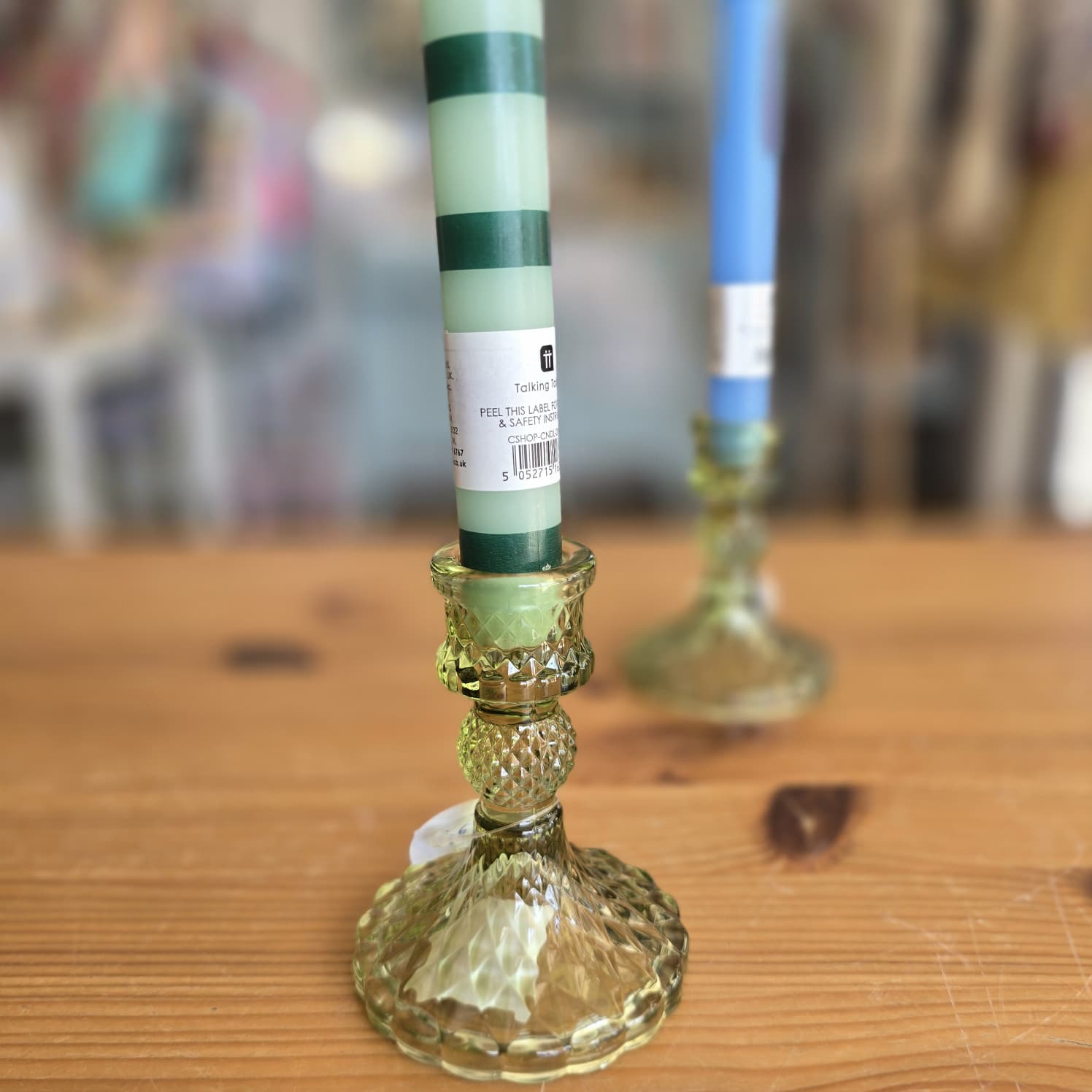 Dois castiçais de vidro verde com velas, uma verde às riscas e outra azul, sobre uma mesa de madeira.