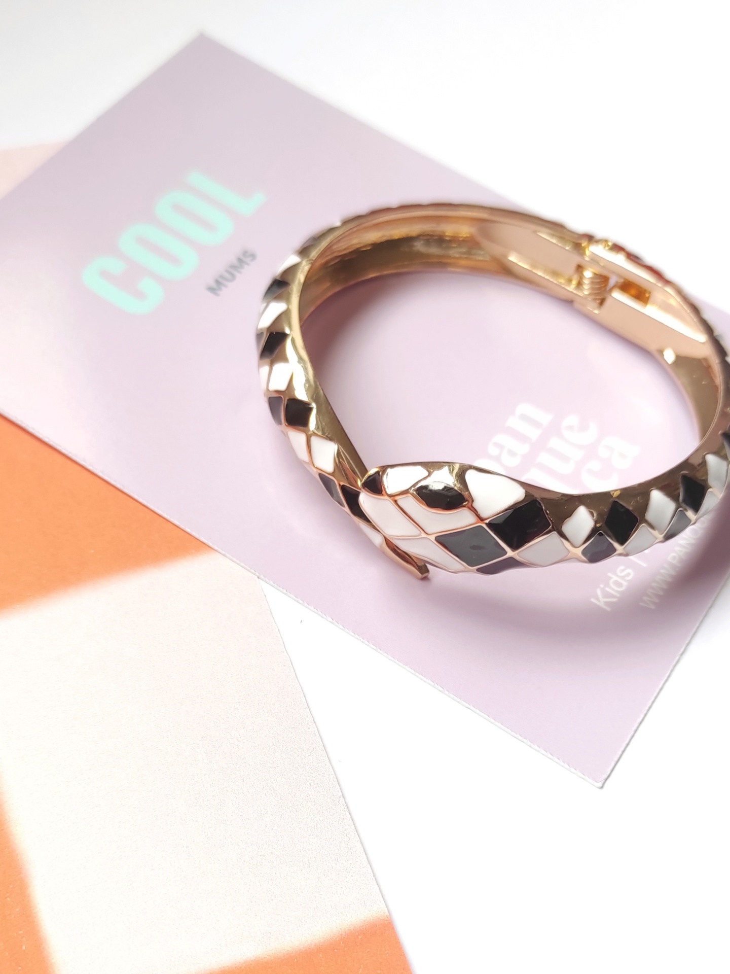 Pulseira dourada com padrão xadrez preto e branco sobre cartão lilás com texto e fundo laranja.