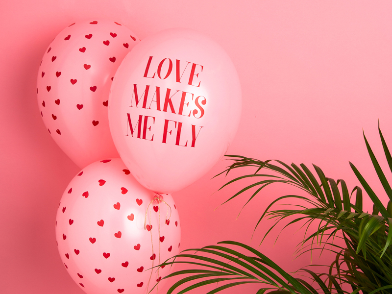 Três balões cor-de-rosa com corações e texto 'LOVE MAKES ME FLY' ao lado de uma planta verde