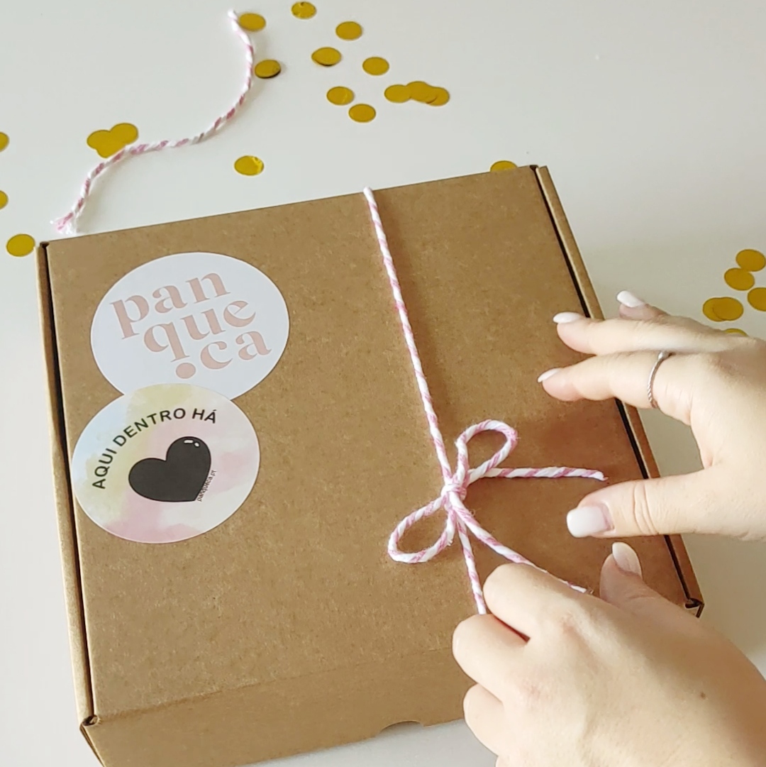 Caixa de cartão castanho com cordão rosa e branco a ser amarrado, etiquetas com texto e confetis ao redor