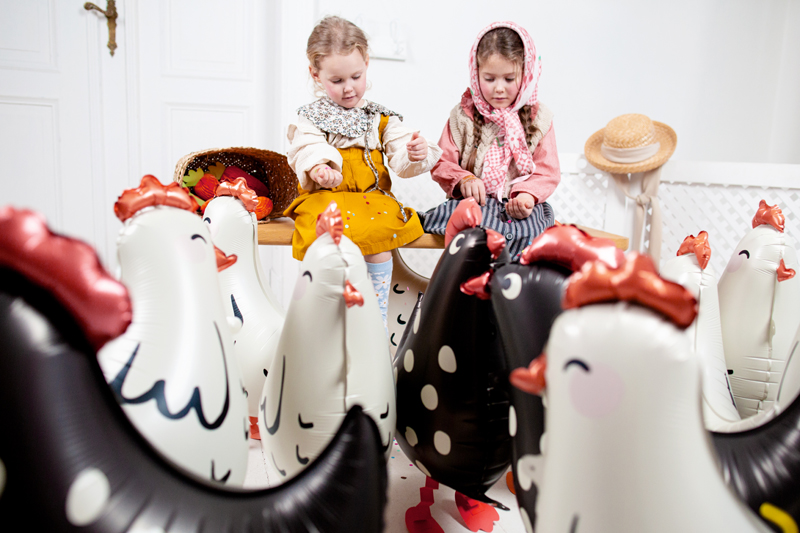 Duas crianças sentadas entre galinhas insufláveis pretas e brancas com detalhes vermelhos num ambiente interior branco