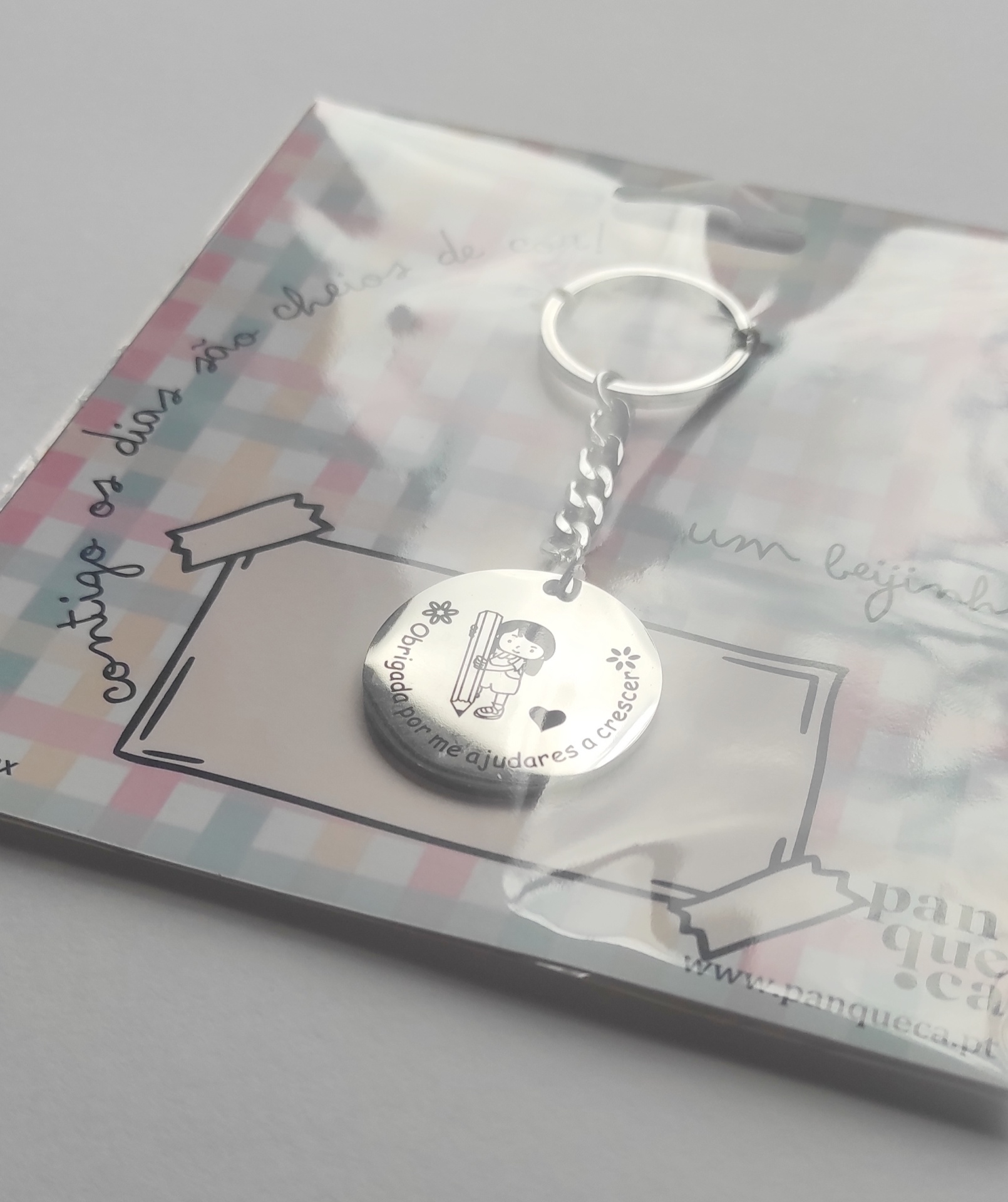 Porta-chaves prateado com pendente redondo e gravação, em embalagem transparente com fundo quadriculado.