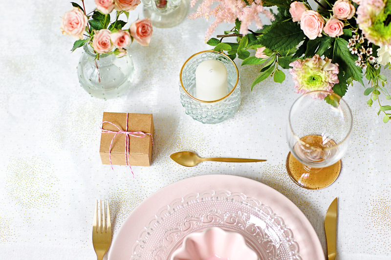 Mesa posta com prato rosa decorado, talheres dourados, copo com bebida, vela e flores
