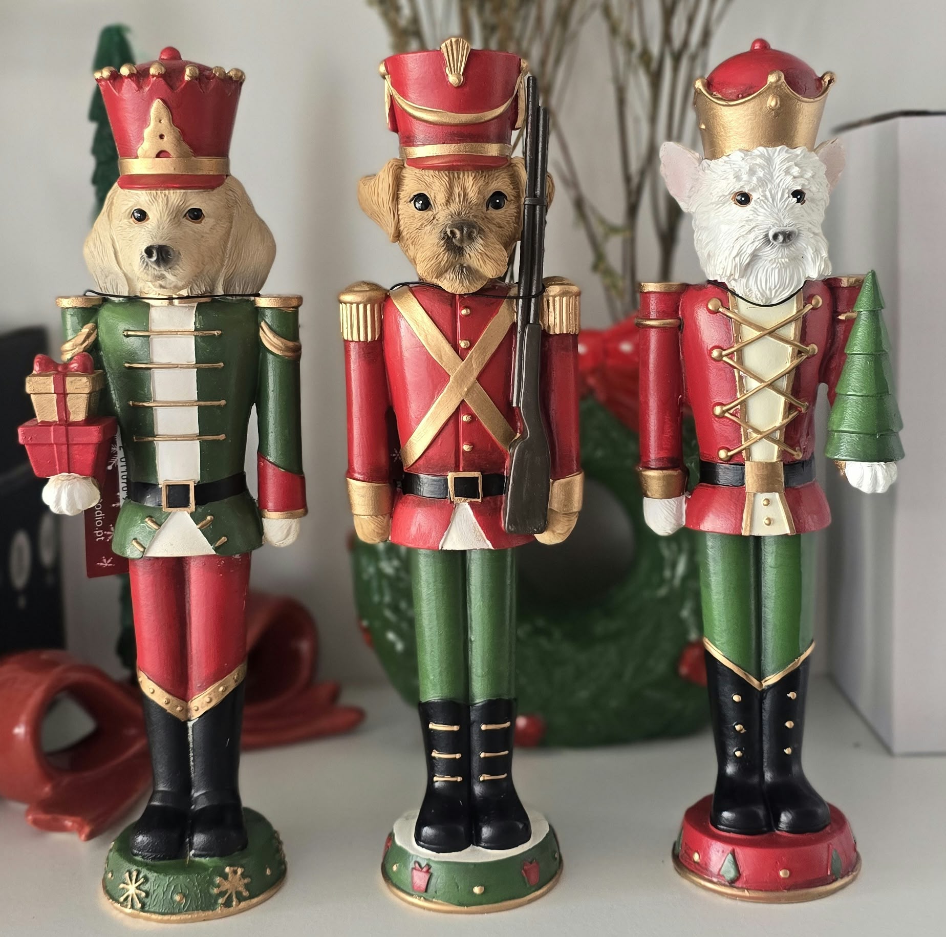 Três figuras de cães como soldados do quebra-nozes vestindo uniformes vermelhos, verdes e dourados, segurando presentes e pinheiro, sobre uma superfície branca