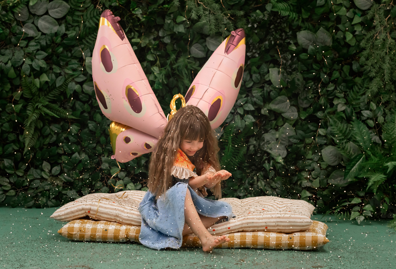 Criança com asas de borboleta rosa sentada em almofadas num chão verde, com fundo de plantas.