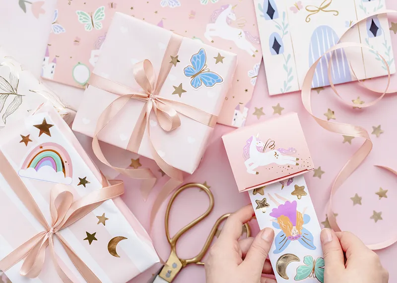Presentes embrulhados em papel rosa com fitas e autocolantes, acompanhados de uma tesoura.