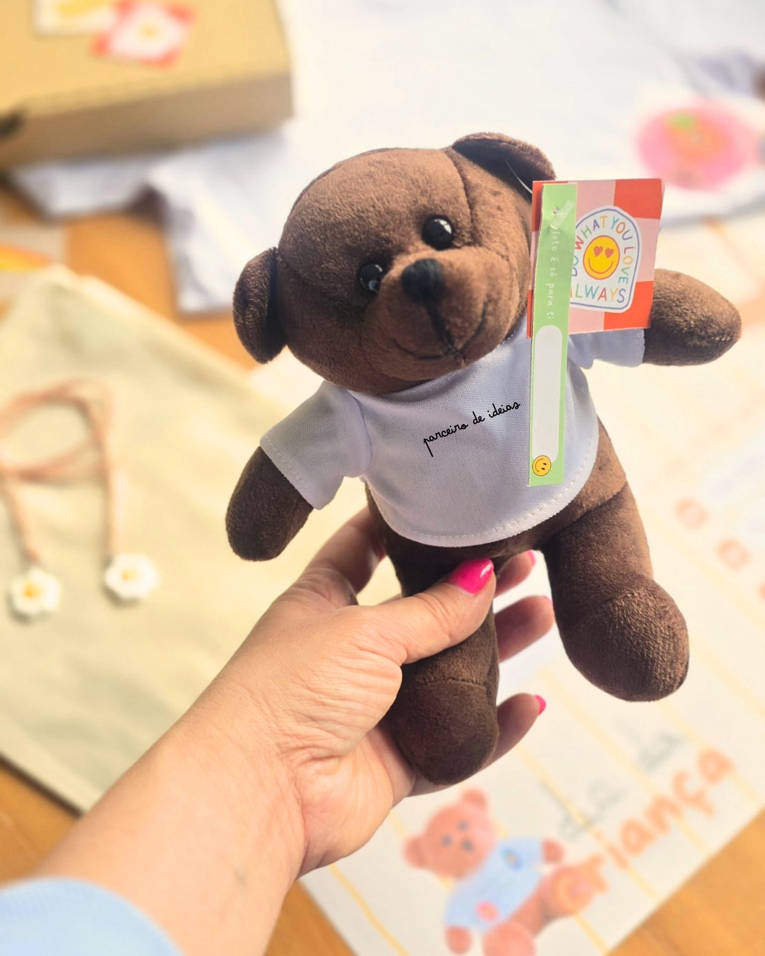 Urso de peluche castanho com t-shirt branca e etiquetas coloridas, segurado por mão com unhas pintadas de rosa.