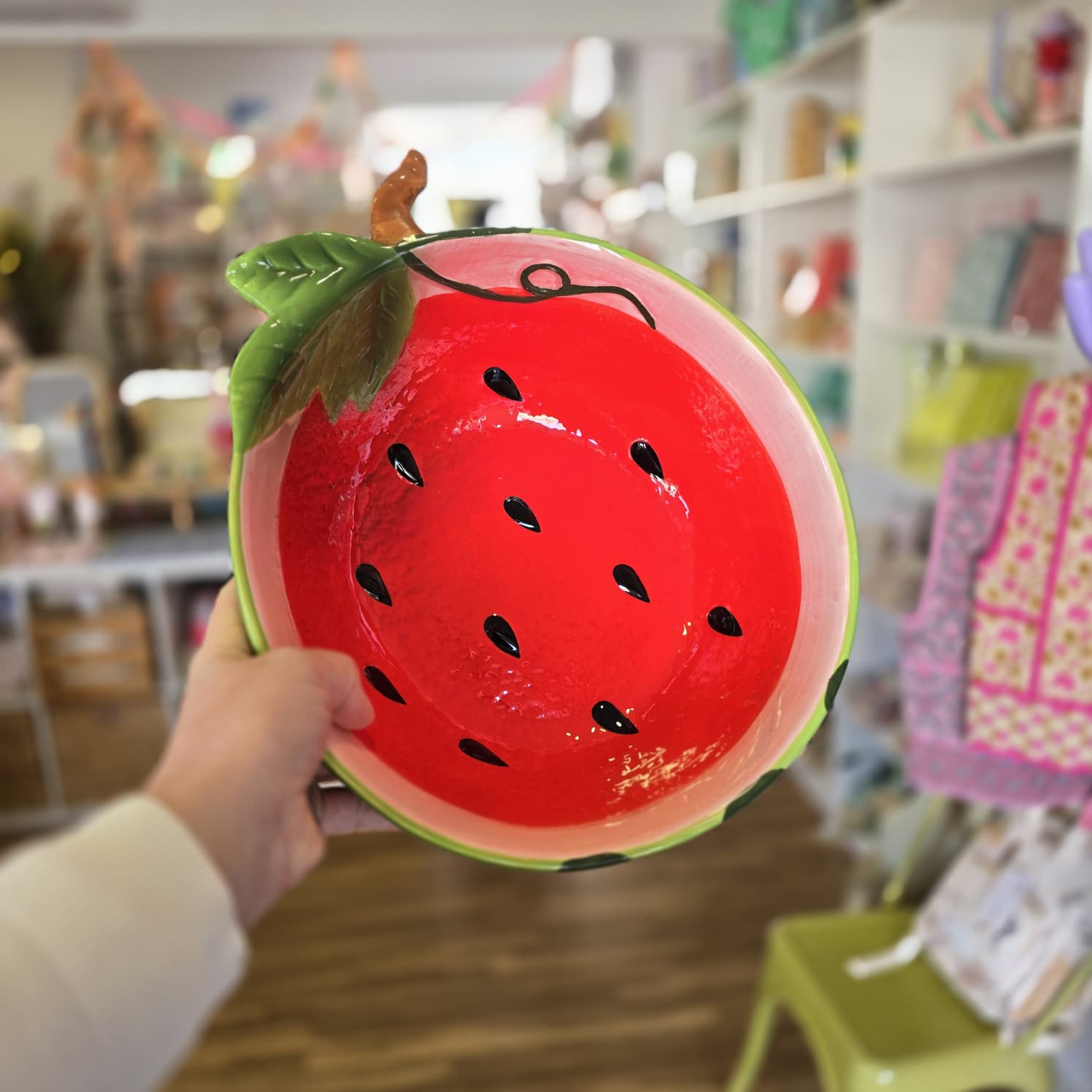 Taça de cerâmica decorada como melancia com sementes e folha, segurada em mão no interior de uma loja.