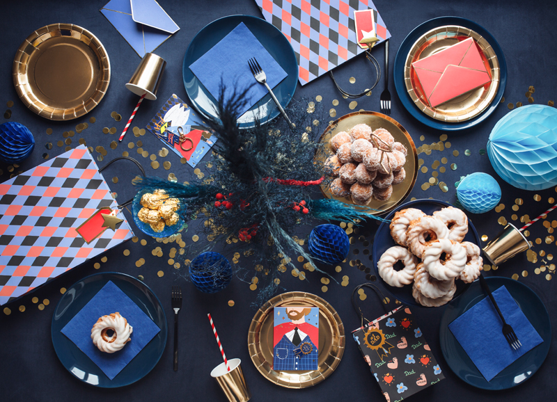 Mesa decorada com pratos, guardanapos, copos dourados e balões azuis para festa.