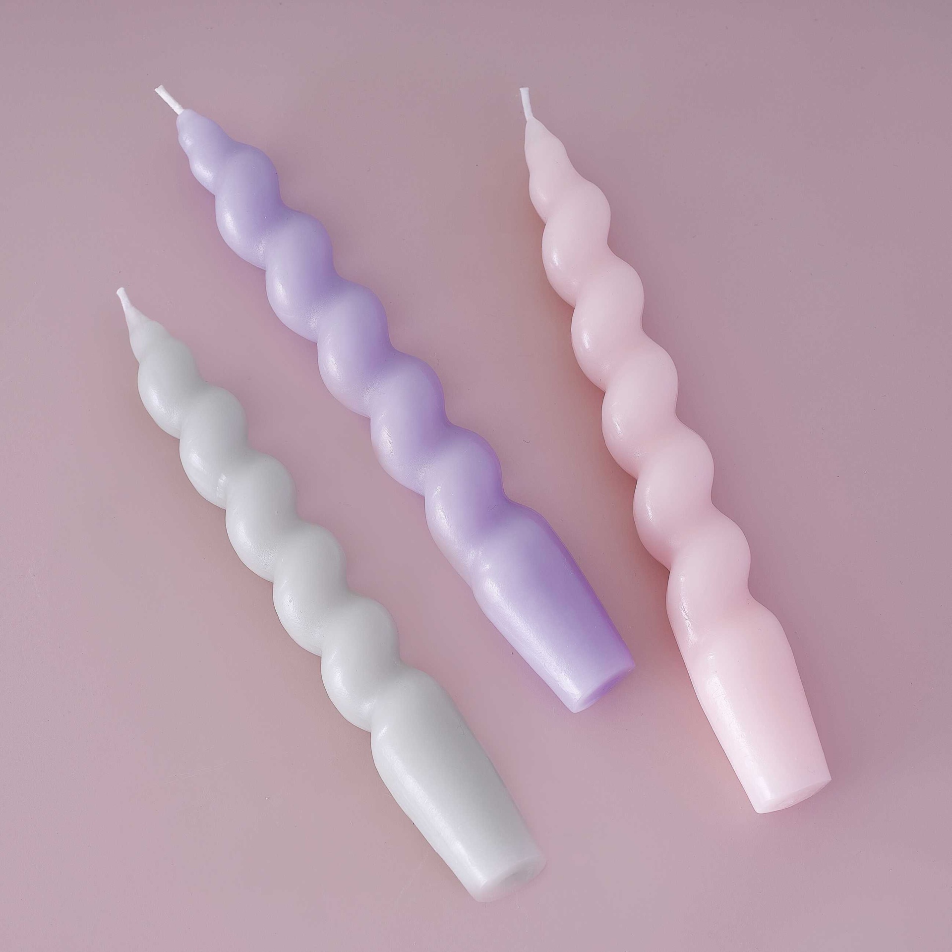Três velas torcidas em branco, lilás e rosa num fundo rosa.