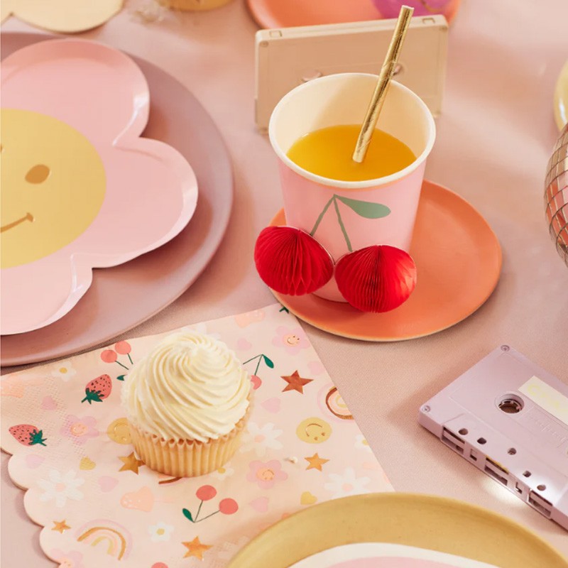 Copo de papel rosa com desenho de cerejas, cupcake e fita cassete cor-de-rosa sobre mesa rosa