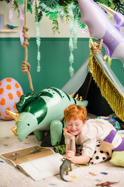 Criança pequena brincando com dinossauro insuflável verde em quarto decorado com planta e balões.