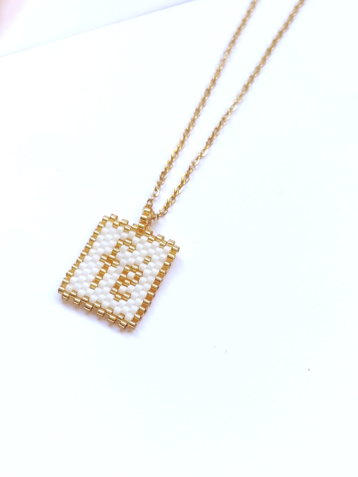 Colar dourado com pingente retangular de missangas brancas e douradas com a letra E