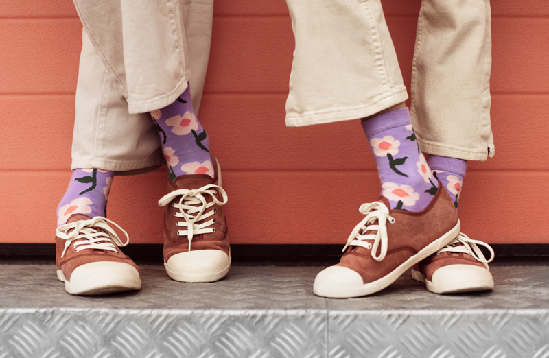 Pessoas calçando sapatilhas castanhas e meias roxas floridas, sentadas junto a parede cor de tijolo