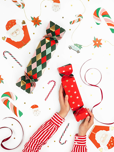 Duas mãos seguram caixa de presente vermelha com estrelas, caixas coloridas e motivos natalícios ao fundo.