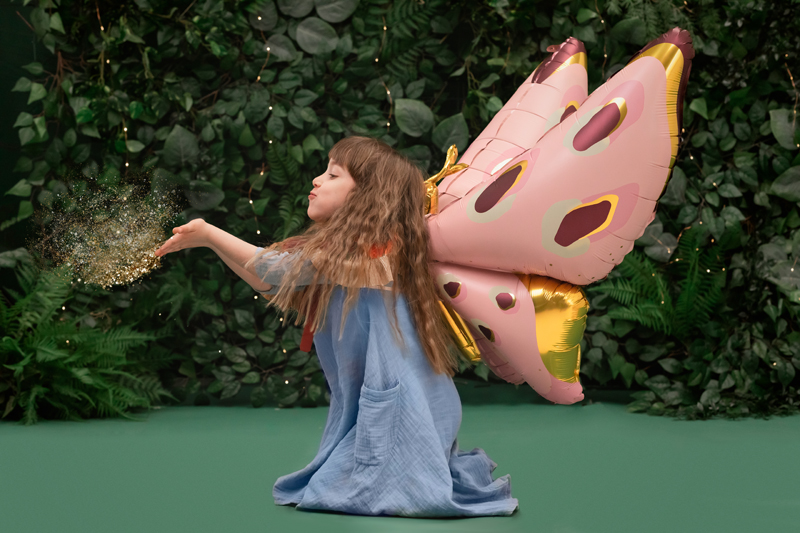 Criança com balões em forma de asas de borboleta cor de rosa e dourado num fundo verde com plantas