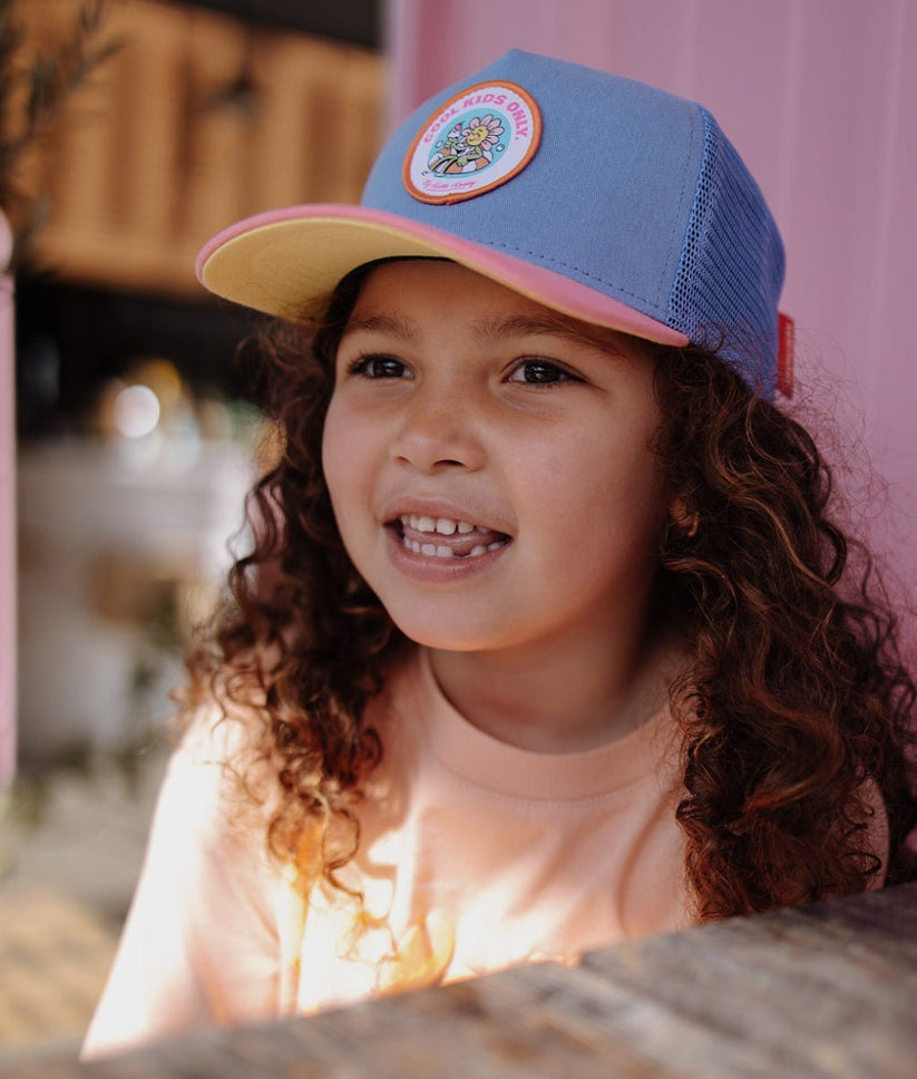 Menina usando boné azul claro, amarelo e rosa com etiqueta redonda com texto e flor.