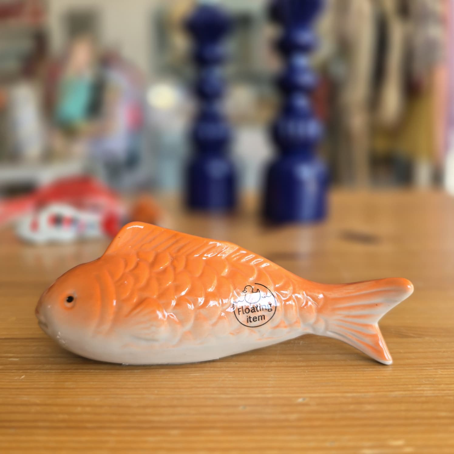 Peixe decorativo de cerâmica laranja e branco em madeira com dois objetos azuis ao fundo.