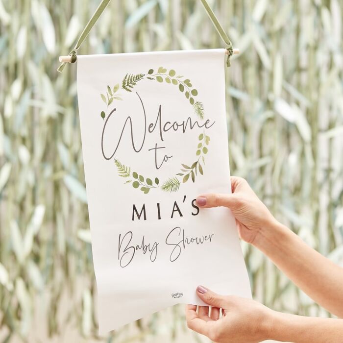 Pendurante de tecido com texto 'Welcome to MIA'S Baby Shower' e moldura de folhas verdes.
