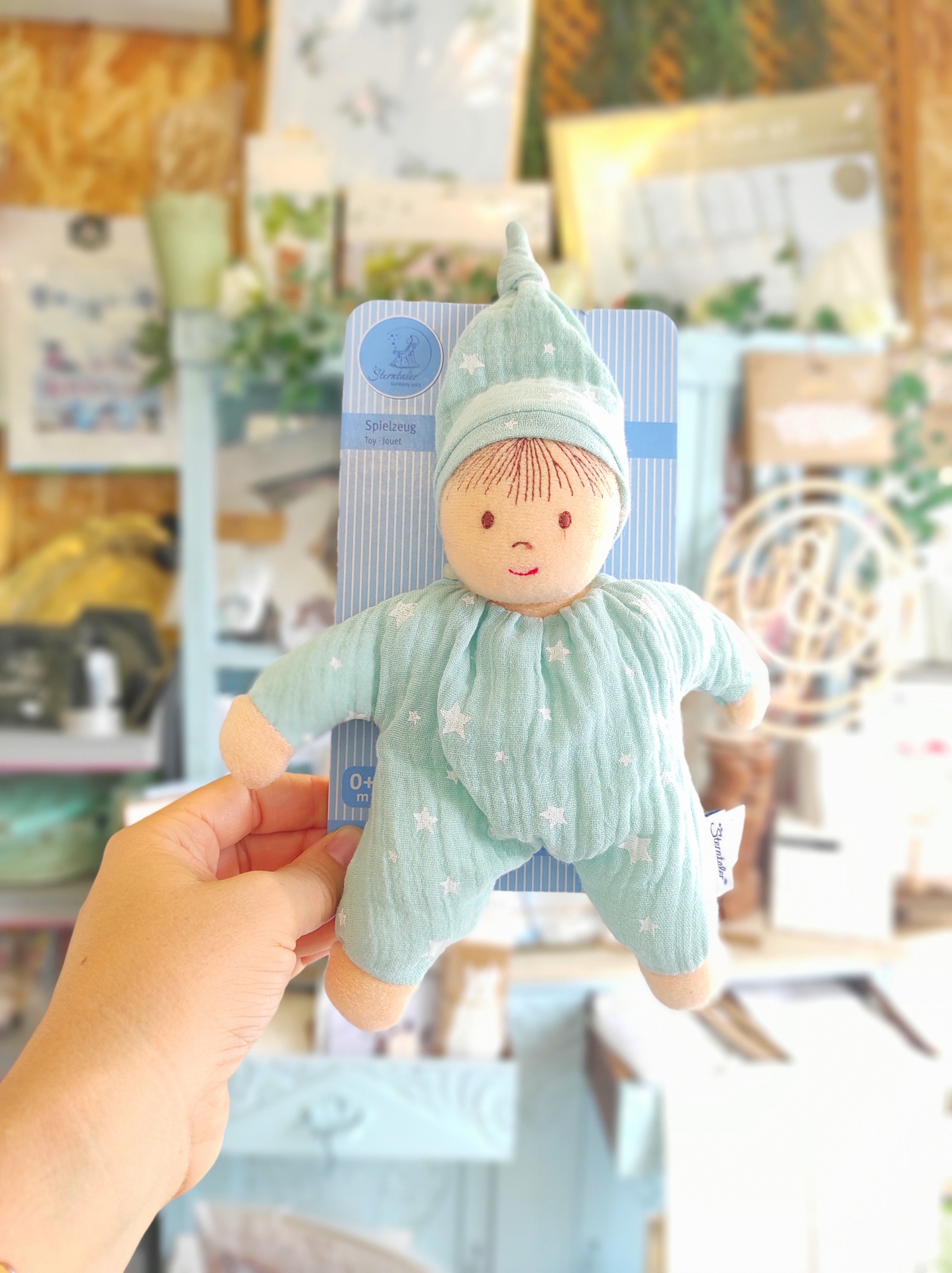 Boneco de tecido verde água com estampado de estrelas e rosto bordado, em embalagem azul e branca