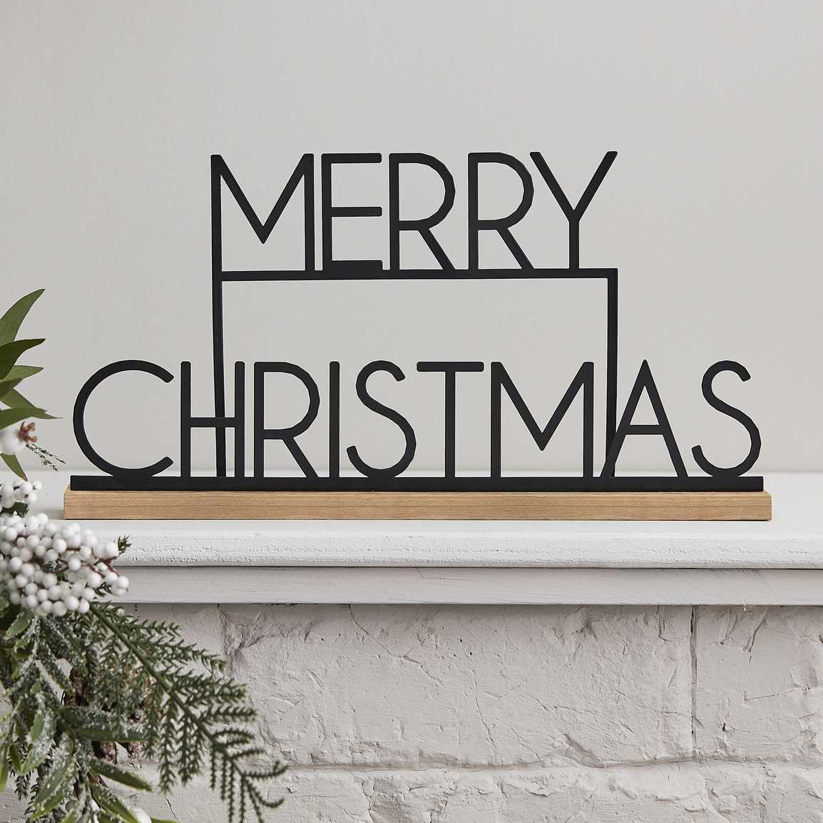 Decoração metálica preta com texto 'MERRY CHRISTMAS' em base de madeira sobre lareira branca
