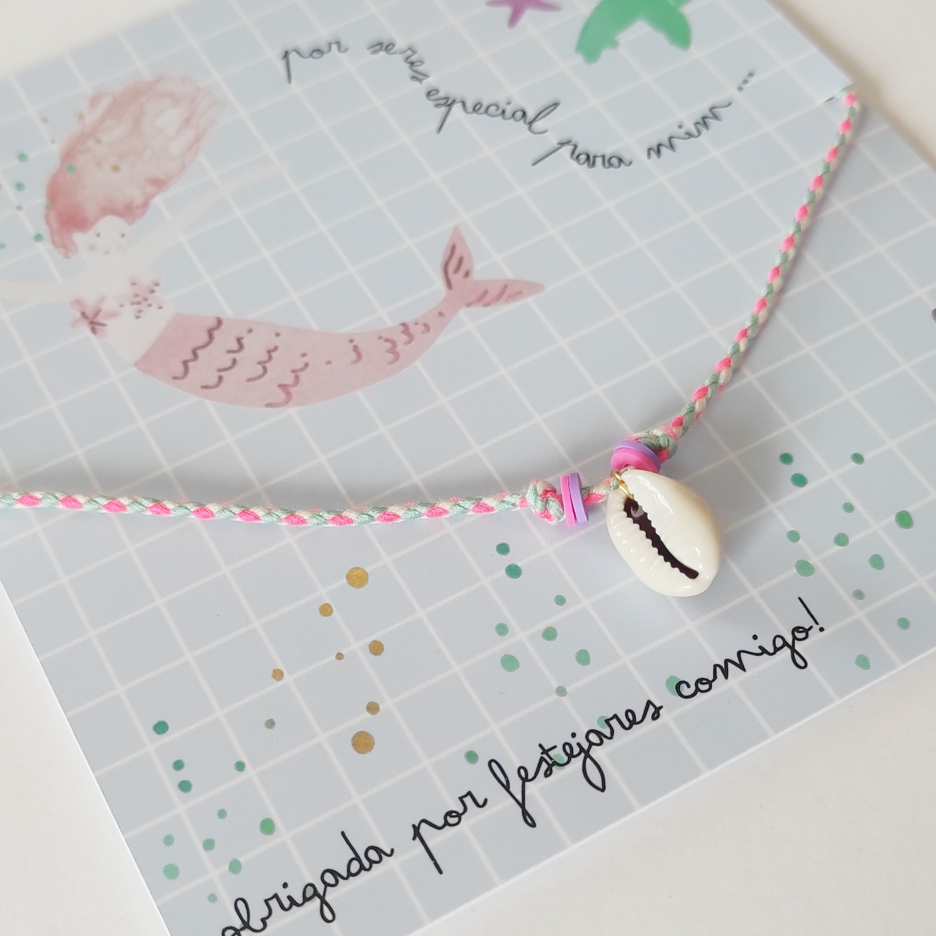 Pulsera de concha blanca con hilo rosa y blanco sobre tarjeta azul con sirena y texto en portugués
