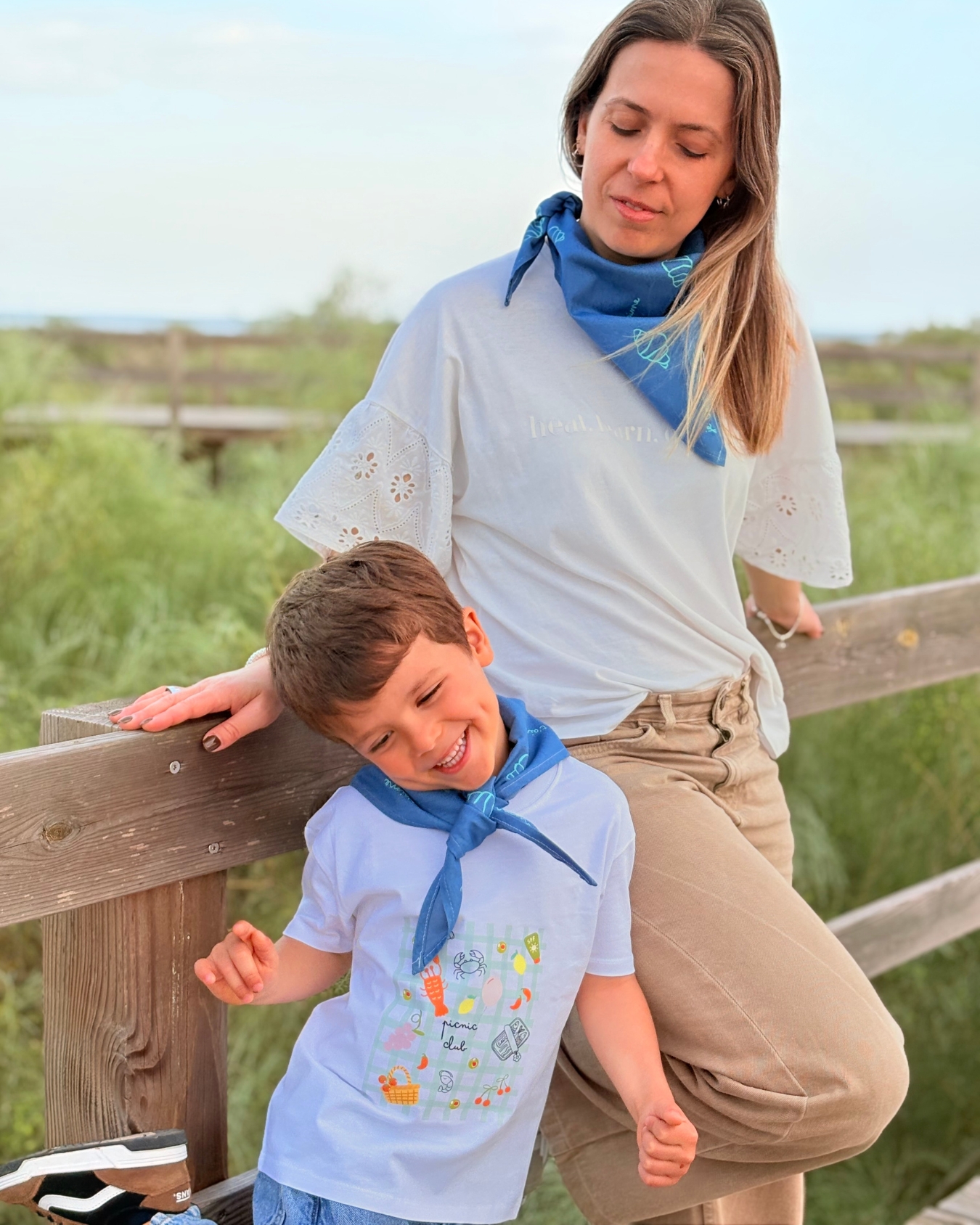 Criança e mulher com t-shirts brancas e bandanas azuis encostados numa vedação de madeira ao ar livre
