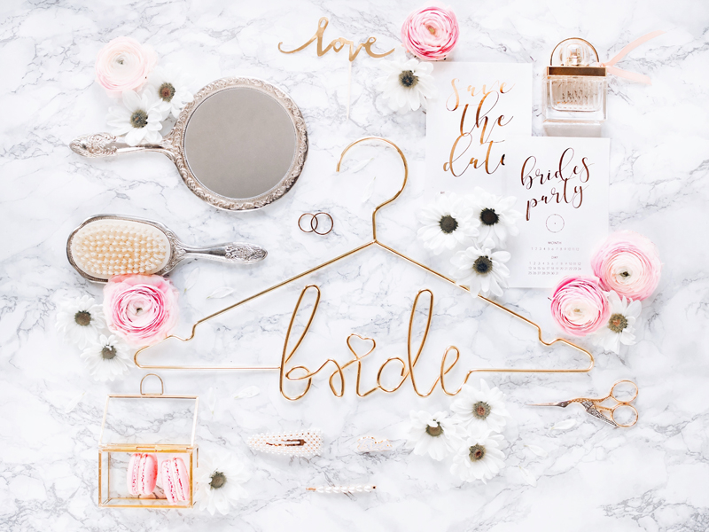 Acessórios de casamento dispostos numa superfície marmoreada branca, incluindo cabide dourado com a palavra 'bride' e convite 'Save the date bride's party'