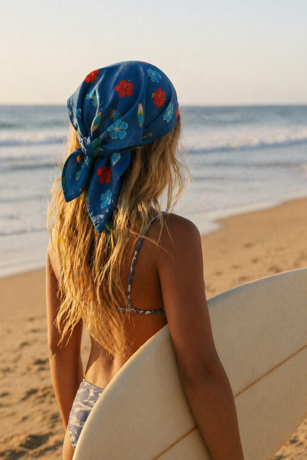 Mulher na praia de costas com lenço azul florido e prancha de surf branca