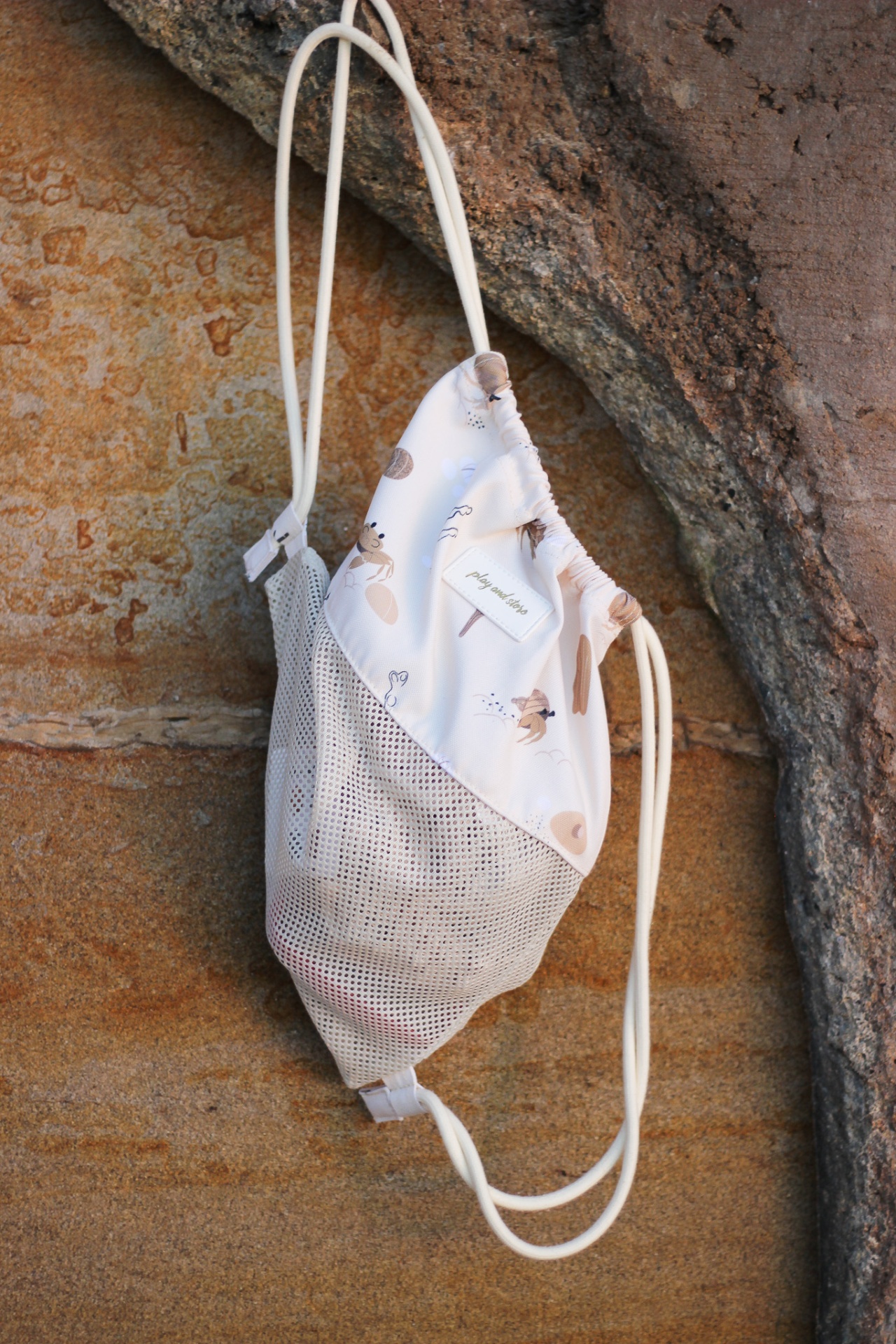 Mochila saco bege claro com padrão e cordas finas pendurada em parede de pedra