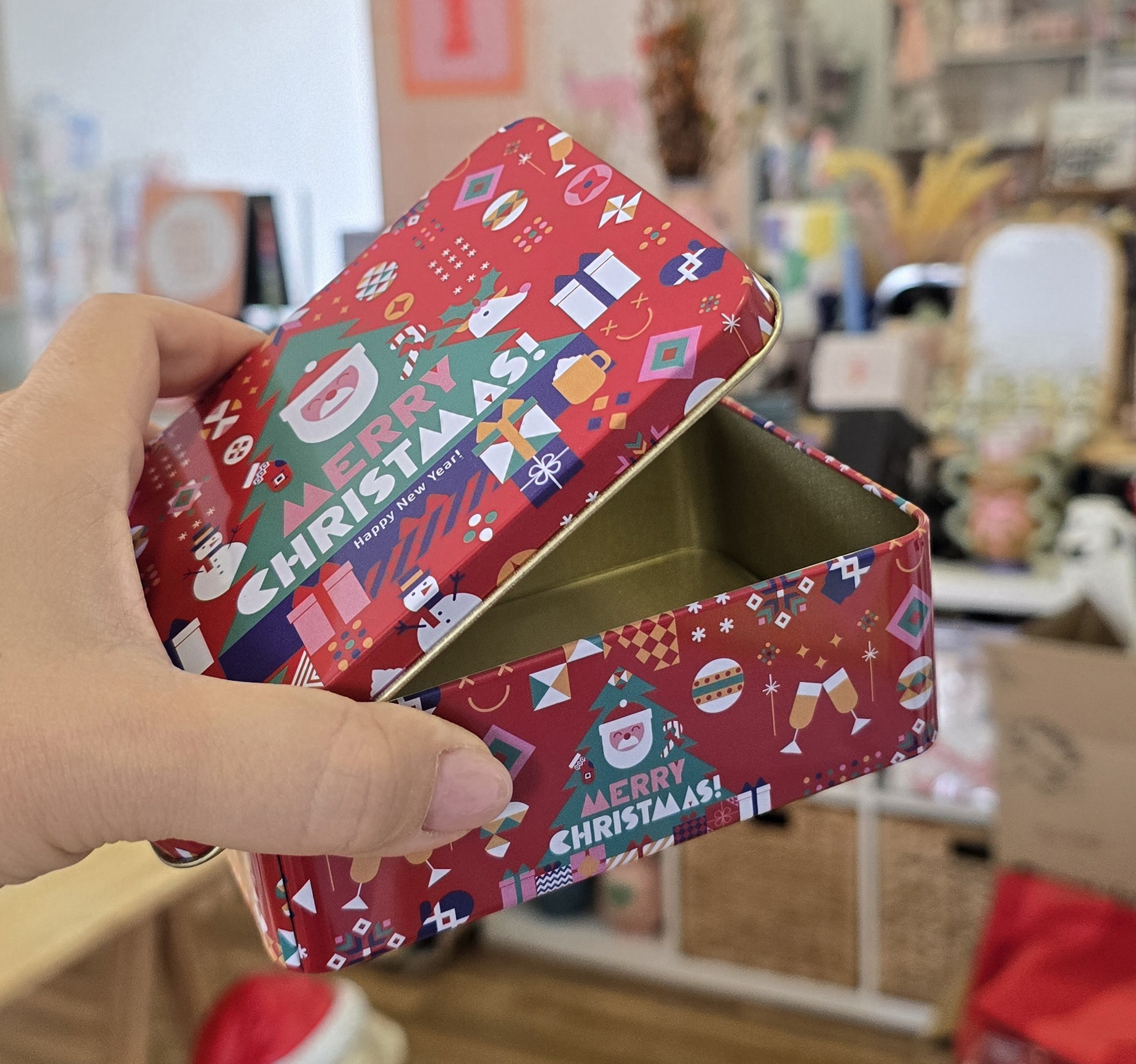Caixa metálica vermelha de Natal com vários ícones coloridos e texto, em ambiente interior.