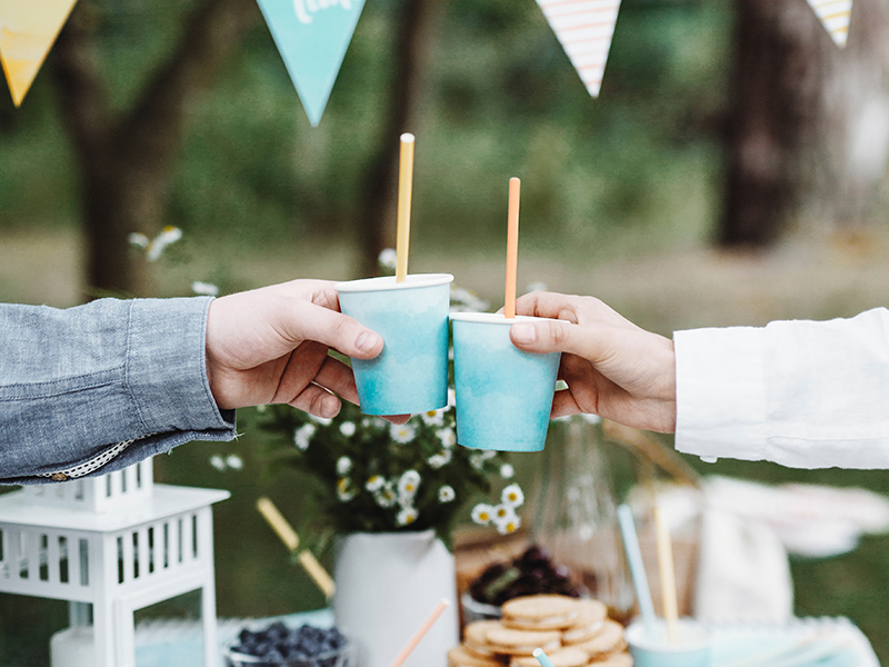 Duas mãos segurando copos de papel azul com palhinhas coloridas ao ar livre com decoração festiva