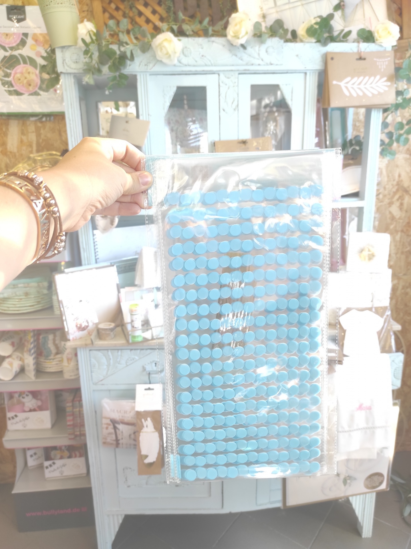 Tapete azul de discos circulares embalado em plástico, segurado por uma mão feminina com pulseiras, fundo com móveis e artigos decorativos.