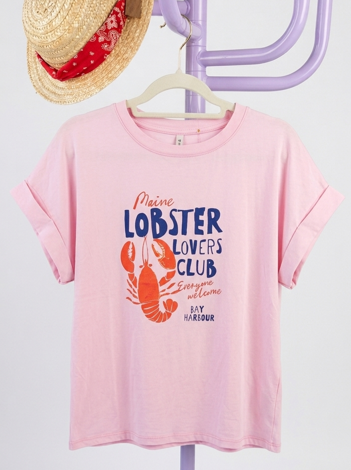 T-shirt rosa com estampa de lagosta e texto, pendurada num cabide creme num suporte lilás com chapéu de palha e lenço vermelho.