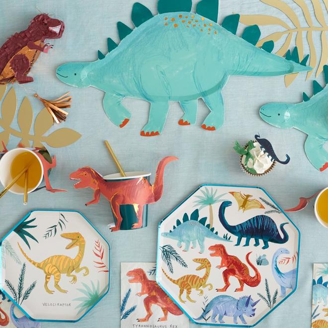 Conjunto de festa com desenhos de dinossauros em pratos e copos sobre toalha azul com padrão de folhas