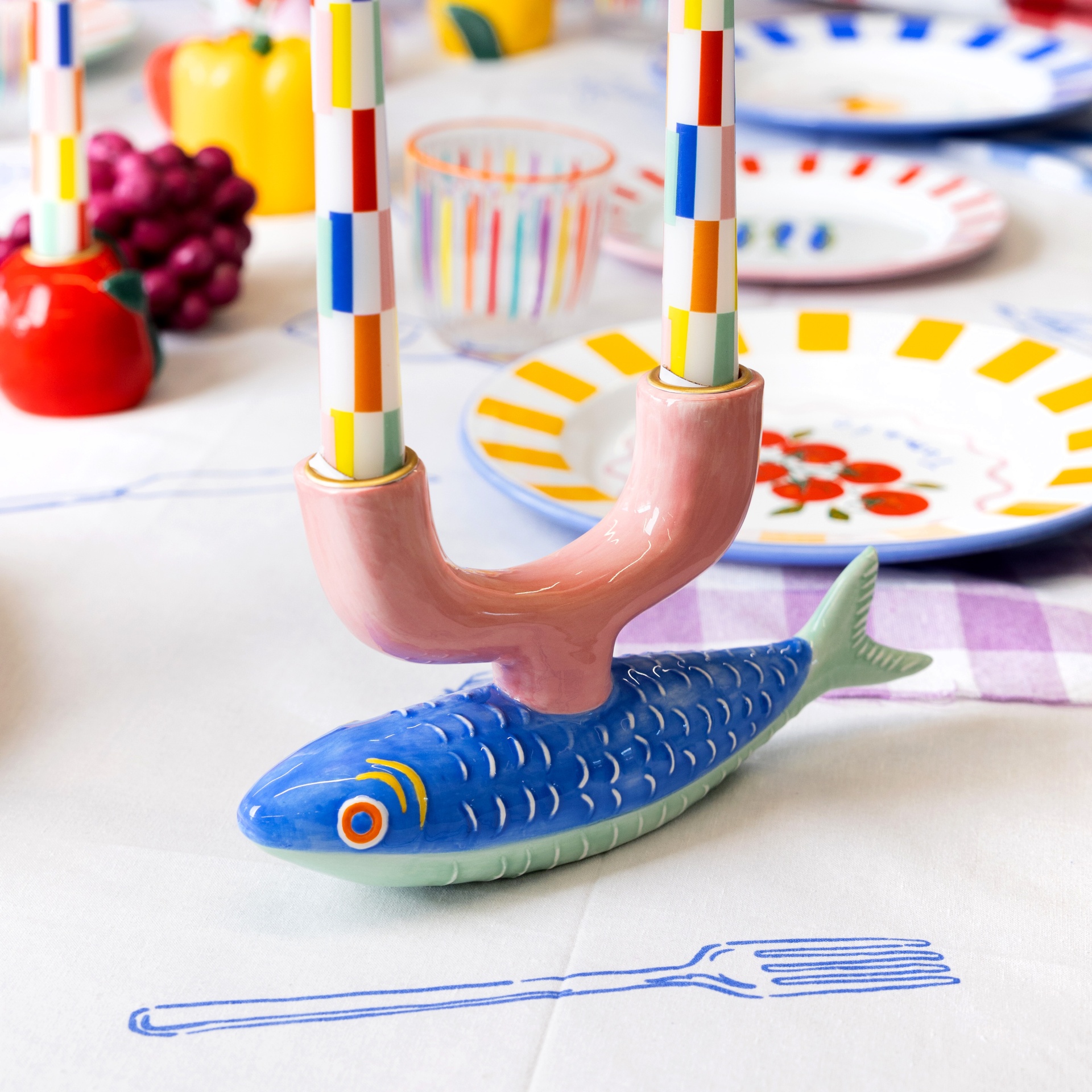 Castiçal cerâmico com base peixe azul, velas riscas coloridas, pratos coloridos numa mesa com toalha branca e desenho azul.