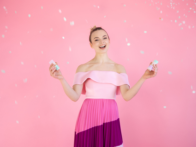 Mulher com vestido rosa e púrpura segurando frascos pequenos em fundo rosa