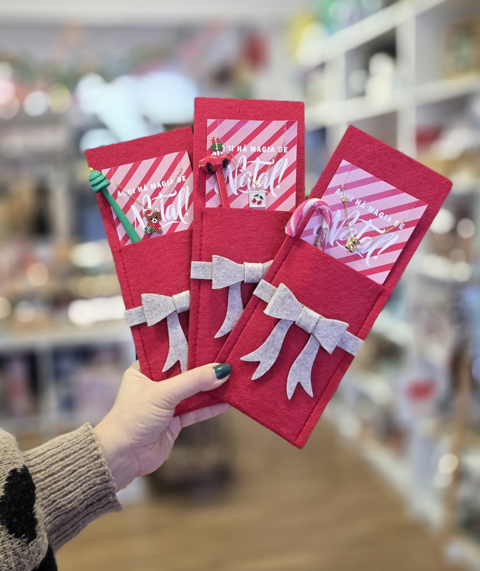Três pegas vermelhas de feltro para telemóvel decoradas com laços e acessórios de Natal em fundo desfocado.
