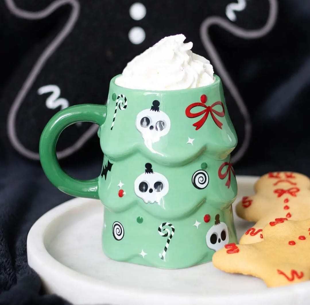 Caneca verde em forma de árvore de Natal decorada com caveiras e doces, com chantilly, em prato com biscoito de gengibre.