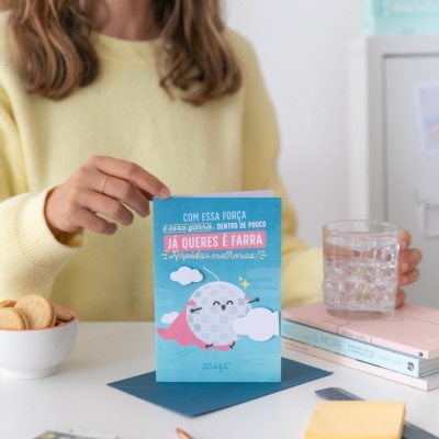 Cartão azul com bola espelhada e capa de super-herói sobre mesa com bolachas e copo de água