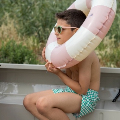 Menino com bóia insuflável branca e rosa, fato de banho xadrez verde e branco sentado numa estrutura metálica