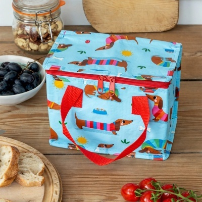 Mala térmica azul com padrão de cães dachshund coloridos e alças vermelhas numa mesa de madeira com comida