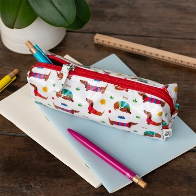 Estojo branco com padrão de cães coloridos e fecho vermelho sobre cadernos e mesa de madeira