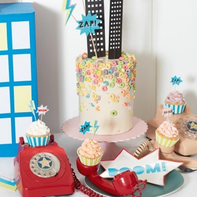 Bolo decorado com cereais coloridos e toppers de prédios e palavras em estilo de quadrinhos, com telefones antigos vermelhos e cupcakes ao redor