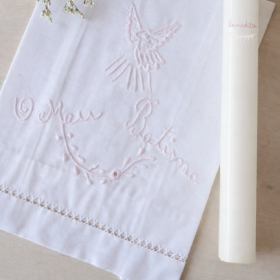 Toalha branca bordada com pomba e texto em rosa junto a uma vela branca com texto benedita