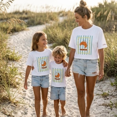 Mulher e duas crianças com t-shirts brancas com croissant, calções jeans e descalços na praia