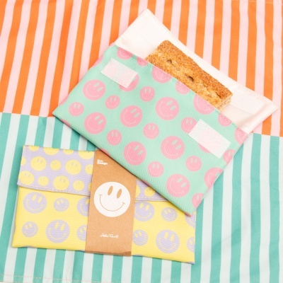 Duas bolsas reutilizáveis para alimentos com padrão de carinhas sorridentes, uma verde com carinhas cor-de-rosa e outra amarela com carinhas lilás, uma com barra de cereal, sobre fundo de riscas coloridas.