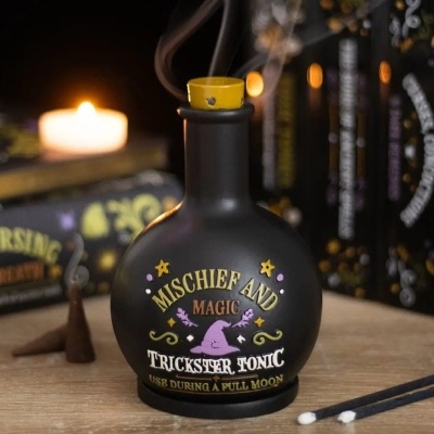 Frasco preto redondo com tampa amarela e rótulo colorido de magia, incenso fumegante e vela acesa num ambiente com livros.
