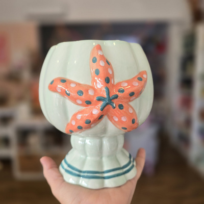 Vaso decorativo branco com estrela do mar em coral e bolinhas, base com riscas azuis, segurado por mão