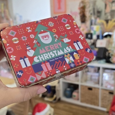 Caixa metálica decorada com motivos natalícios coloridos e texto Merry Christmas segurada por mão